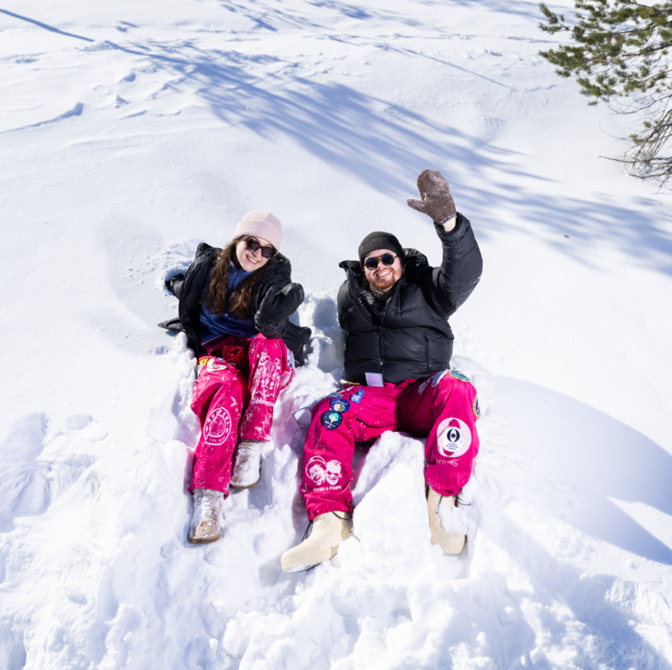 Two students smile and sit on a bed of snow. The other student waves at the camera.