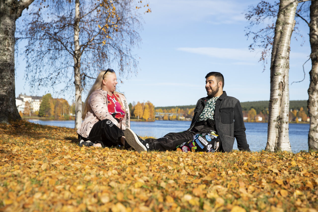 Kaksi yhteiskuntatiedeiden opiskelijaa opiskelijahaalarit päällä istuskelemassa ja juttelemassa Kemijoen rannalla syysauringon paisteessa.