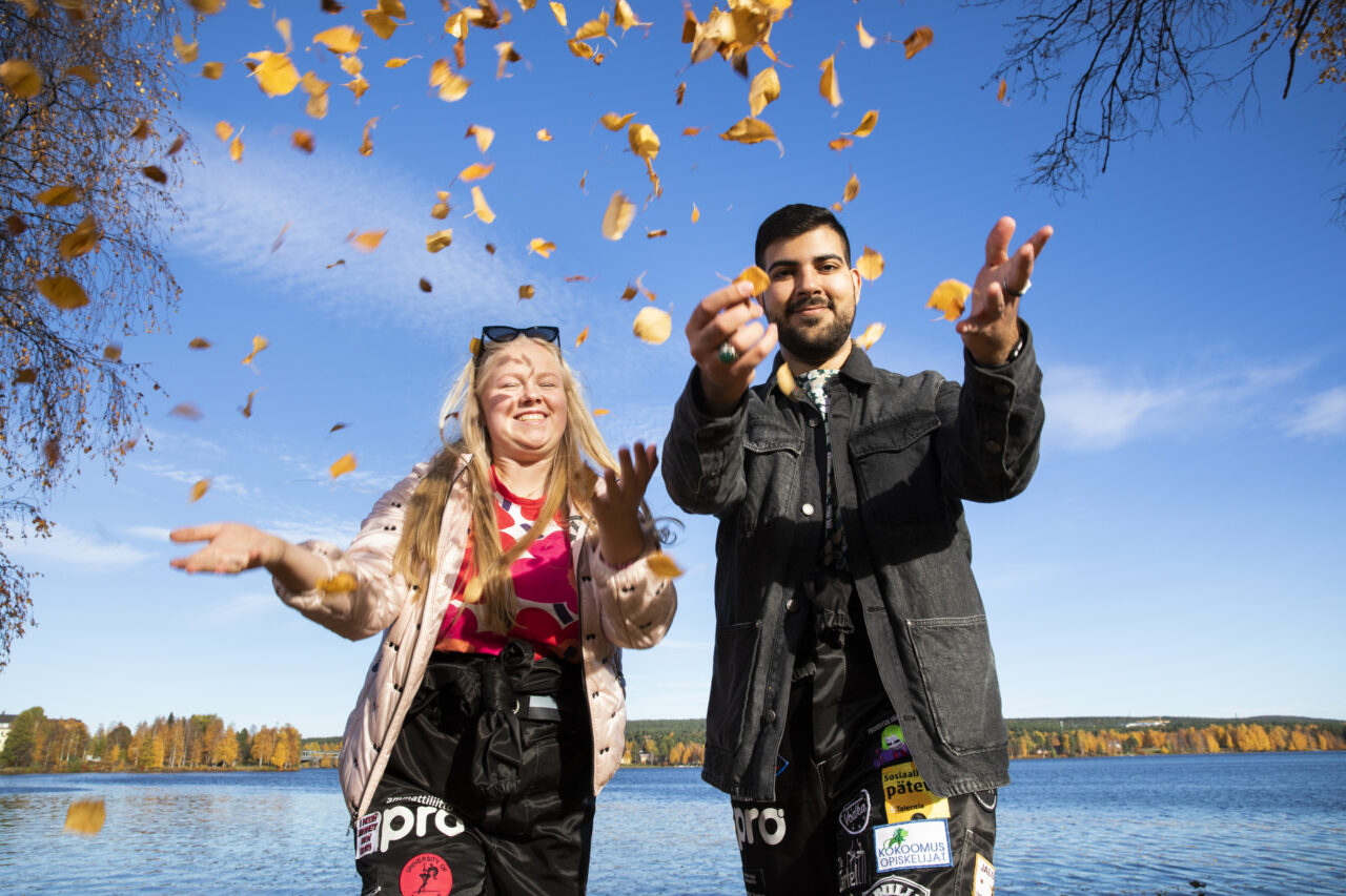 Two smiling students throw yellow birch tree leaves at the camera. An autumnal river landscape can be seen in the background.