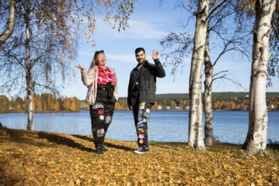 Kaksi yhteiskuntatiedeiden opiskelijaa opiskelijahaalarit päällä vilkuttamassa kameralle Kemijoen rannalla syysauringon paisteessa.