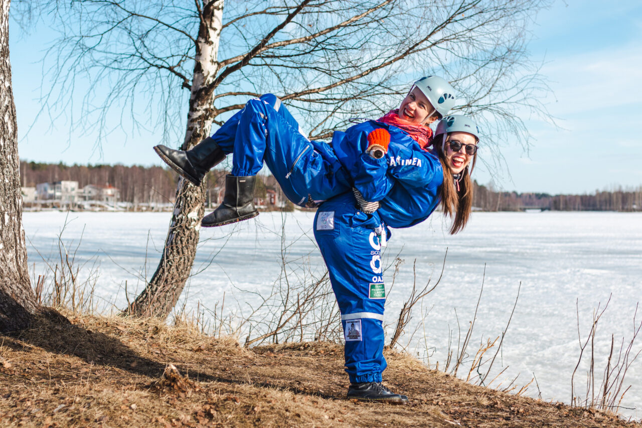Kaksi kestävyyskasvatuksen opiskelijaa opiskelijahaalarit päällä Kemijoen rannalla.