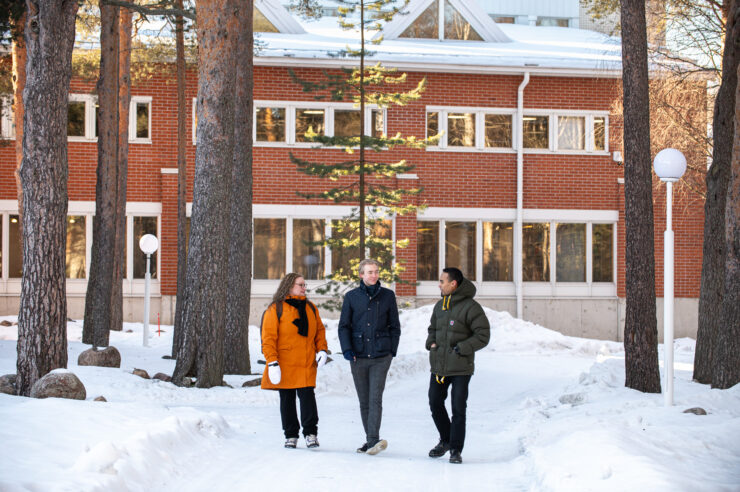 "Kolme opiskelijaa kävelevät talvella yliopistorakennuksen edessä mäntymetsässä."